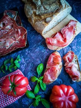 Ham, tomato, bread and herbs in composition on dark background with rough texture