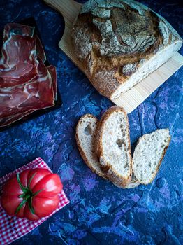 Ham, tomato, bread and herbs in composition on dark background with rough texture
