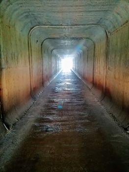 Square tunnel passing under a highway with sunlight entering at the end of the tunnel