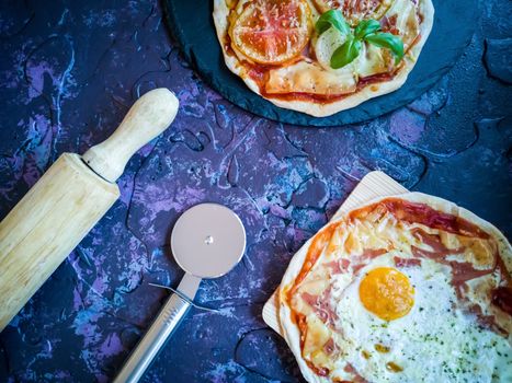 Homemade pizzas and utensils on dark background in lilac tones