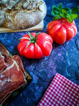 Ham, tomato, bread and herbs in composition on dark background with rough texture