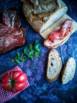 Ham, tomato, bread and herbs in composition on dark background with rough texture