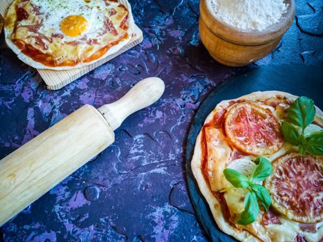 Homemade pizzas and utensils on dark background in lilac tones