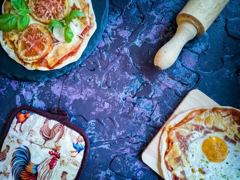 Homemade pizzas and utensils on dark background in lilac tones