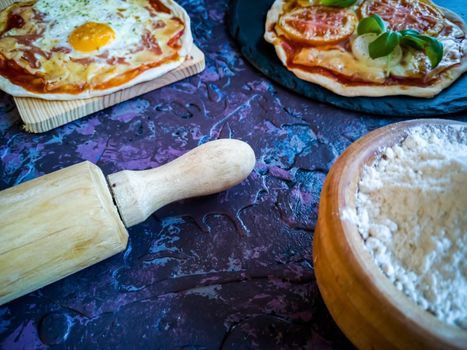 Homemade pizzas and utensils on dark background in lilac tones
