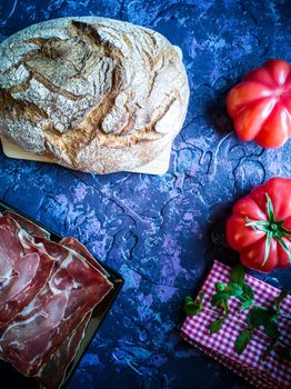 Ham, tomato, bread and herbs in composition on dark background with rough texture