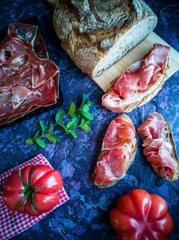 Ham, tomato, bread and herbs in composition on dark background with rough texture