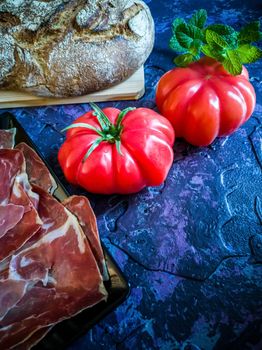 Ham, tomato, bread and herbs in composition on dark background with rough texture