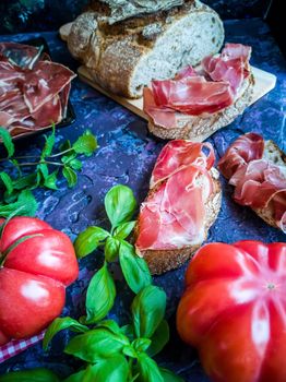 Ham, tomato, bread and herbs in composition on dark background with rough texture
