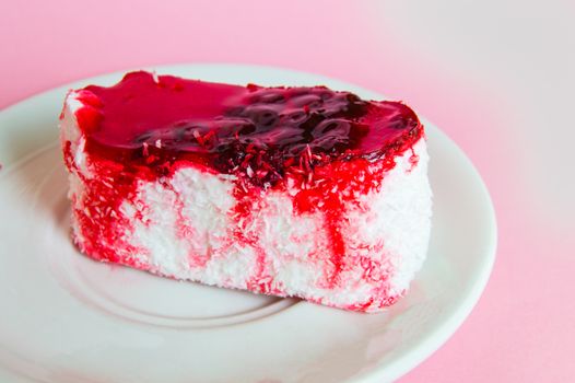 Exquisite souffle cake with lingonberry jelly, with berry glaze and coconut on a white plate, top view close-up, pink background.