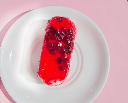 Exquisite souffle cake with lingonberry jelly, with berry glaze and coconut on a white plate, top view close-up, pink background.