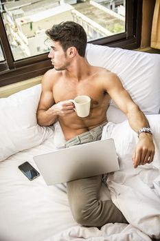 Naked young man with muscular body on bed with mug or cup in hand with coffee or tea, looking at laptop computer screen