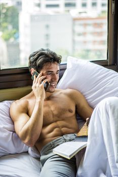 Sexy handsome young man laying shirtless on his bed next to window, holding a coffee or tea cup while reading a book