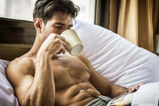 Sexy handsome young man laying shirtless on his bed next to window, holding a coffee or tea cup while reading a book