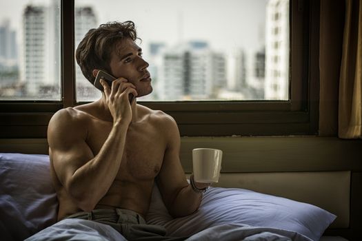 Naked young man with muscular body on bed with mug or cup in hand with coffee or tea talking on cell phone