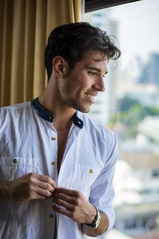 Sexy handsome young man standing and dressing, in his bedroom next to window curtains