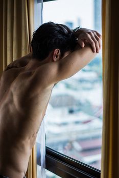 Sexy sad young man standing shirtless in his bedroom next to window curtains, looking out