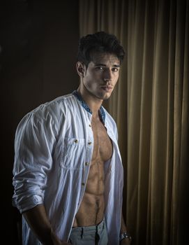 Sexy handsome young man standing and dressing, in his bedroom next to window curtains, looking at camera with shirt open on naked muscular torso
