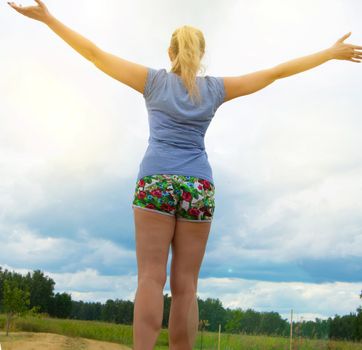 Happy young blonde wearing shorts and a t-shirt stands on the road against the sky with her arms outstretched, illuminated by sunlight on a summer day.
