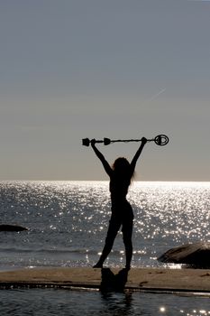 silhouette of a girl on the sand of a beach with a metal detector in her hands raised above her head. Against the shimmering glare of the sun on the surface of the sea. Counter shooting