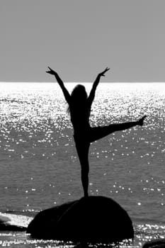 silhouette of a girl standing on a stone in the sea with raised arms and leg. Against the shimmering glare of the sun on the surface of the sea. Counter shooting.