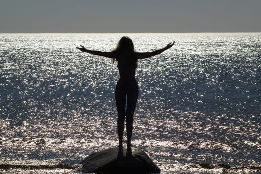 Photo of a girl standing on a stone with her arms spread apart. By the sea. Against the shimmering glare of the sun on the surface of the sea. Counter shooting.