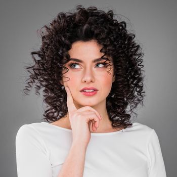 Close up portrait of pretty confident thoughtful girl, holding hand near the face, looking seriously up, standing over grey background