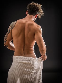 Portrait of naked handsome young man with languishing look covering crotch with a towel, against black background