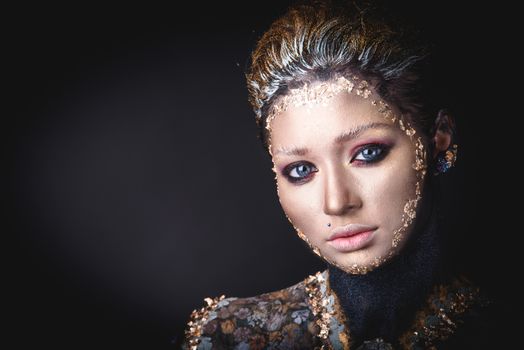 Portrait of a blue eyed young girl with creative golden makeup in the style of icon painting isolated on black background