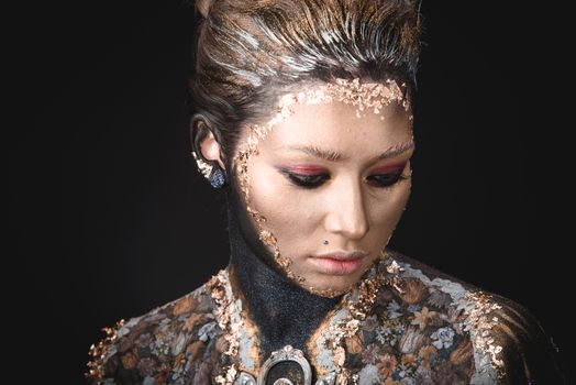 Portrait of a blue eyed young girl with creative golden makeup in the style of icon painting isolated on black background