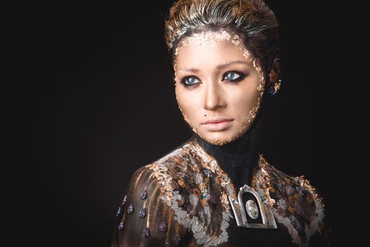 Portrait of a blue eyed young girl with creative golden makeup in the style of icon painting isolated on black background