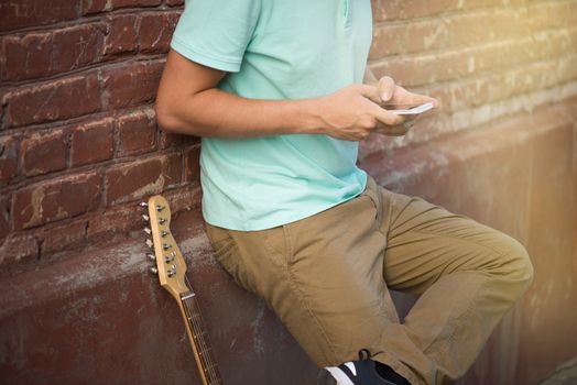 Summer, party, young stylish guy standing near brown brick wall, hands holding mobile phone next to guitar, sunlight.