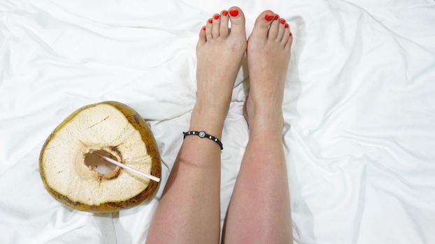 Female bare feet on a white sheet, top view, closeup. Coconut cocktail near the feet. Hotel by the sea concept