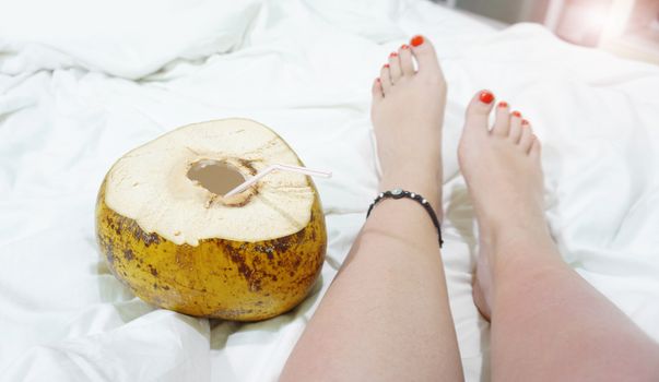 Female bare feet on a white sheet, top view, closeup. Coconut cocktail near the feet. Hotel by the sea concept