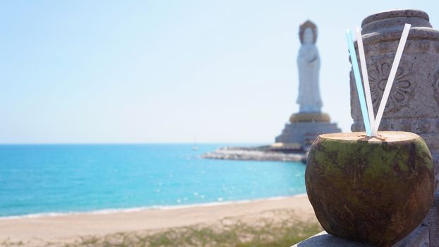 Guanyin of Nanshan, the white statue of the bodhisattva Guanyi near the Nanshan Temple of Sanya, China. In the foreground a coconut cocktail, the sea