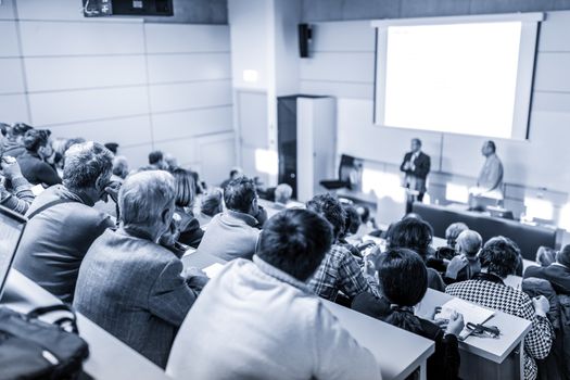 Speaker giving a talk in conference hall at business event. Audience at the conference hall. Business and Entrepreneurship concept. Focus on unrecognizable people in audience. Blue toned.