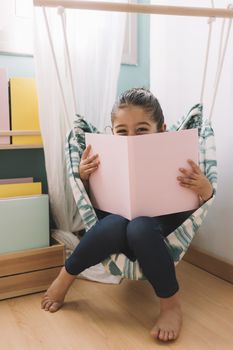 vertical photo of a sweet happy little girl reading a book in her room near the window, funny lovely child having fun in kids room