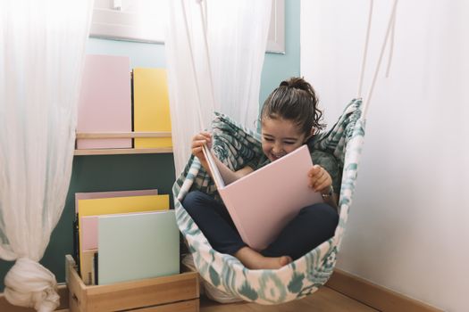 little girl laughing while reading a book in her hammock near the window, funny lovely child having fun in her kids room