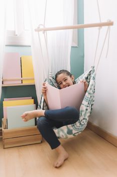 vertical photo of a little girl laughing while reading a book in her kids room near the window, funny lovely child having fun at home, copy space for text