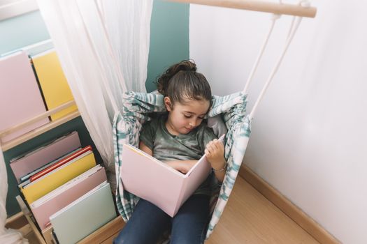 little girl concentrated reading a book at home near the window, funny lovely child having fun in her kids room, copy space for text