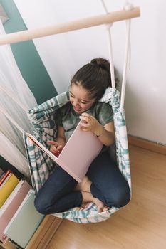 little girl laughs out loud reading a book at home near the window, funny lovely child having fun in her kids room, copy space for text, vertical photo