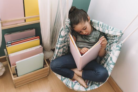 little girl concentrated reading a book in her kids room near the window, funny lovely child having fun at home, copy space for text