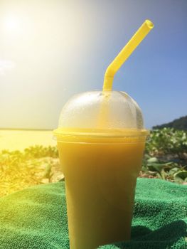 A glass of summer tropical cold cocktail with a misted lid and a straw stands on the beach on a towel against the blue sky and sunlight,