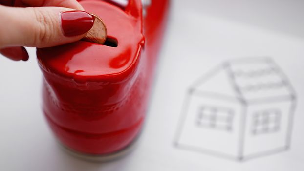 Hand dropping coin into the moneybox. Drawing a house on a white sheet near with a box