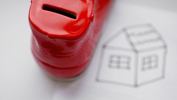 Hand dropping coin into the moneybox. Drawing a house on a white sheet near with a box