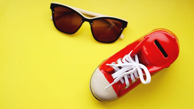 Moneybox in the form of sneakers. Sunglasses on a yellow background. The concept of savings on vacation and summer holidays. Money save