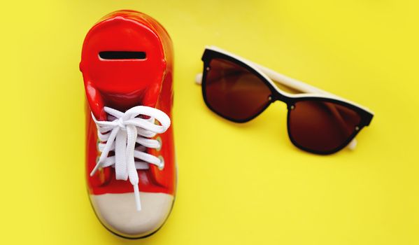 Moneybox in the form of sneakers. Sunglasses on a yellow background. The concept of savings on vacation and summer holidays. Money save