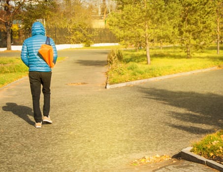 Young man in blue jacket with sports bag, walking in Park, rear view, on Sunny summer day.