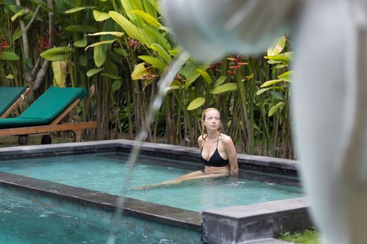 Sensual young woman relaxing in outdoor spa infinity swimming pool surrounded with lush tropical greenery of Ubud, Bali. Wellness, natural beauty and body care concept.