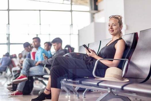 Casual sporty young blond female traveler using her cell phone while waiting to board a plane at the departure gates at the asian airport terminal.